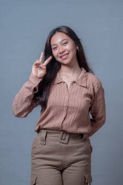 Uzun siyah saçlı, dokulu kahverengi gömlekli gülümseyen Endonezyalı kadın sağ eliyle barış işareti yapıyor. Sade gri bir stüdyo geçmişine karşı neşeyle poz veriyor..
