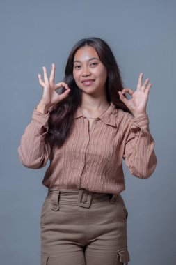 Endonezyalı kadın iki eliyle olumlu bir jest yaparak tarafsız gri bir arkaplana karşı gülerek 