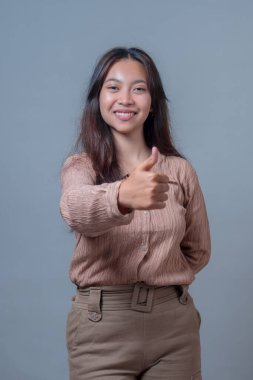 Endonezyalı kadın, sağ eliyle baş parmak işareti yaparken gülümsüyor, günlük giysiler içinde. Tarafsız gri bir arkaplana karşı duran kendine güvenen ve pozitif bir ifadesi var..