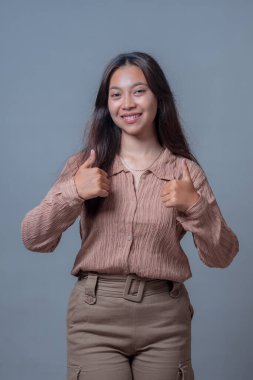 Endonezyalı kadın gülümsüyor ve kendinden emin ve arkadaş canlısı bir ifadeyle başparmağını kaldırıyor, günlük kıyafetler giyiyor ve rahat bir poz veriyor, tarafsız gri bir arka plana karşı..