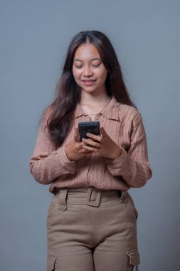 Endonezyalı genç bir kadın akıllı telefon kullanıyor, teknolojiyle iletişim kurarken gülümsüyor. Bej bir bluz ve pantolon giyip modern yaşam tarzını ve aletlerle olan bağlantısını sergiliyor..