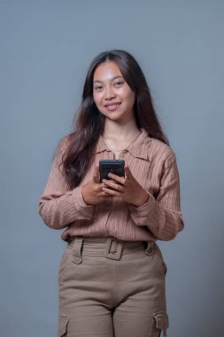 Endonezyalı kadın akıllı telefon kullanıyor, aletle konuşurken gülümsüyor. Bej bir bluz ve pantolon giyerek, modern bir bağlantı ve teknoloji yaşam tarzını somutlaştırıyor..