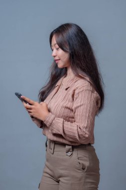 Endonezyalı kadın akıllı telefon kullanıyor. Tarafsız gri bir arkaplanda dikilirken ekrana odaklanmış. Bej bir bluz ve pantolon giyiyor.