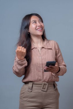 Asyalı kadın yukarı bakarken gülümsüyor, tek eliyle akıllı telefonu tutuyor ve başarılı bir yumruk hareketi yapıyor. Teknoloji ve neşeli dijital yaşam tarzı kavramı.