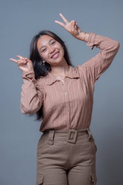 Neşeli bir Endonezyalı kadın iki eliyle barış işareti gösterirken sıcak bir şekilde gülümsüyor. Onun neşeli ifadesi ve neşeli jesti pozitifliği, mutluluğu ve kendine güveni iletir..