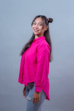 Gülümseyen Asyalı genç bir Endonezyalı kadın portresi, yarı çörek saç stili, canlı pembe bir gömlek ve yırtık kot pantolon giyiyor, tarafsız gri bir arka planı olan güvenli bir şekilde stüdyoda poz veriyor..