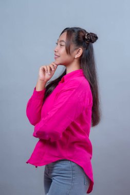 Parlak pembe gömlekli ve yırtık kotlu gülümseyen Endonezyalı bir kadının profili. Stüdyo ortamında elleri çenesinde gri bir arka planda duruyor..