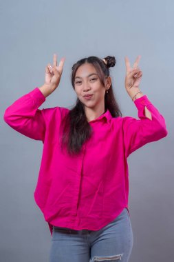 Kendine güvenen genç Endonezyalı kadın canlı pembe bir gömlek ve yırtık kot pantolon giyiyor, neşeli bir surat ifadesiyle gülümsüyor ve profesyonel bir stüdyoda çift barış işareti gösteriyor..