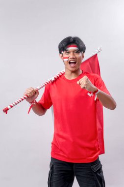 Endonezya 'nın Bağımsızlık Günü' nü tutkuyla kutlayan genç bir adam vatansever yüz boyalı kırmızı bir tişört giyiyor. Omzunda Endonezya bayrağıyla enerjik bir şekilde poz veriyor. Gurur ve coşkuyu ifade ediyor.