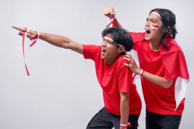 Kırmızı tişörtlü iki hevesli genç, kırmızı-beyaz saç bantları ve yüz boyası takarak, güçlü ulusal gururlarını göstererek ileri doğru işaret ederken tutkuyla tezahürat yapıyorlar..