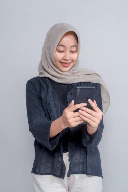 Bej tesettürlü ve koyu renk elbiseli gülümseyen genç Asyalı kadın iki eliyle akıllı telefonuna bakıyor, gri arka planda duruyor, dijital yaşam tarzını temsil ediyor..