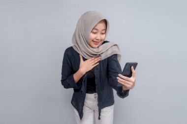Asyalı bir kadın tesettürlü akıllı telefona bakıyor sıcak bir gülümsemeyle, elleri göğsünde minnettar veya duygusal bir jest yapıyor. Takdir kavramı, dijital iletişim, ya da iyi haber.