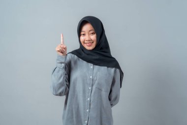 Siyah tesettürlü ve gri tişörtlü gülümseyen genç bir müslüman kadının portresi, tek parmakla yukarıya doğru, düz gri bir arkaplan üzerinde duruyor..