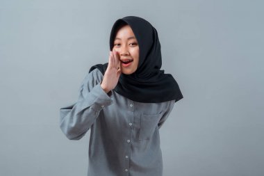 Siyah tesettürlü ve gri gömlekli genç Müslüman bir kadın elini ağzına atıyor, sanki fısıldıyor ya da anons ediyor gibi, arka planda neşeli bir ifadeyle..