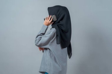 Siyah tesettürlü, gri tişörtlü, Asyalı genç bir Müslüman kadının telefon görüşmesi yaparken sade bir arkaplanda duruşu, sakin ve odaklanmış görünümü..