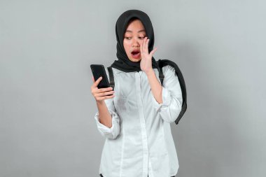 Siyah tesettürlü ve beyaz tişörtlü Asyalı genç kız, akıllı telefonundan okula dijital heyecanı simgeleyen bir şey okurken şaşırmış görünüyor..