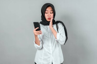 Başörtüsü ve sırt çantası takan genç Müslüman kız öğrenci, akıllı telefonunu kontrol ederken şaşırmış görünüyor. Beklenmedik haberlere tepki veriyor, gri arka planda okula dönmeye hazır..