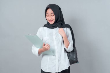 Siyah tesettür ve beyaz gömlek giyen genç Müslüman bir kadın, bir pano okurken ve bir sırt çantası tutarken, eğitimi, odaklanmayı ve geri dönüş ruhunu temsil ederken gülümsüyor..