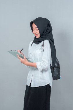 Siyah tesettürlü ve beyaz tişörtlü gülümseyen Müslüman öğrenci bir panoya odaklanmış ve öğrenmeye hazır. Yeni akademik hedefleri simgeleyen bir sırt çantası takıyor..