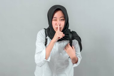 Tesettürlü Asyalı kız öğrenci sessiz bir jest yapıyor, işaret parmağı dudaklarında, beyaz gömlek ve sırt çantası giyiyor, neşeli bir şekilde göz kırpıyor, gri arka planda izole edilmiş, stüdyo portresi.