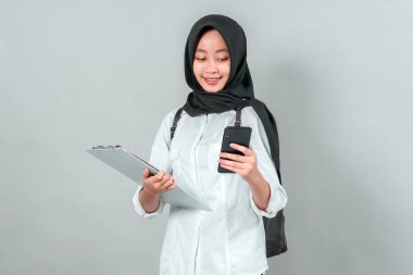 Mutlu Müslüman kız öğrenci tesettüre ve sırt çantasına sarılmış, elinde dosya ve akıllı telefon, telefonuna bakarken gülümsüyor, yeni akademik öğretim yılına hazır..