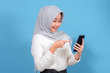 Açık gri tesettür ve beyaz süveter giyen neşeli, genç bir Asyalı kadın, mavi arka planda ışıl ışıl gülümseyen akıllı telefon ekranını işaret ediyor..