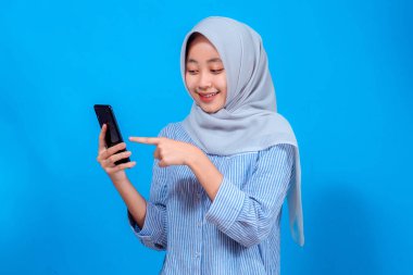 Gri tesettürlü ve çizgili tişörtlü neşeli Asyalı genç Müslüman kadın akıllı telefon ekranını gösterirken gülümsüyor, mutlu ve heyecanlı bir yüz ifadesi sergiliyor..