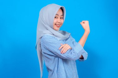 Kendine güvenen, tesettürlü, genç, Asyalı Müslüman bir kadın. Kollarını kasarken gururla gülümsüyor. Gücünü, gücünü ve pozitifliğini mavi arka planda gösteriyor..