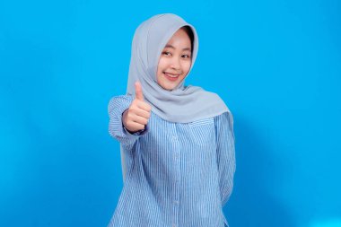 Açık gri tesettürlü ve mavi çizgili tişörtlü neşeli genç bir kadın ışıl ışıl gülümsüyor ve canlı mavi bir arkaplanda kendine güvenen bir jest yapıyor..