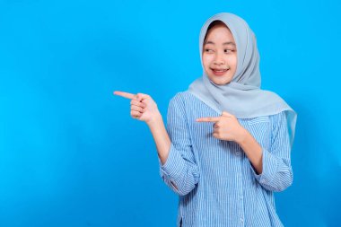 Gri tesettürlü ve çizgili tişörtlü gülümseyen genç Müslüman kadın iki eliyle neşeyle kenarı gösteriyor, heyecan, coşku ve pozitif merak ifade ediyor..