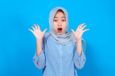 Genç Asyalı kadın tesettür ve çizgili gömlek giyiyor. Şaşırmış bir ifade sergiliyor. Ağzı açık ve mavi bir arkaplanda el kaldırıyor..