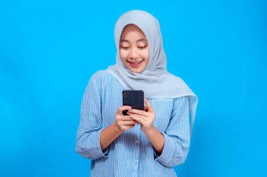 Açık gri tesettürlü ve çizgili tişörtlü genç bir Asyalı kadın akıllı telefonuna bakarken sıcacık gülümsüyor, canlı mavi bir arka planda duruyor..