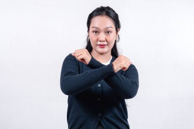 Siyah hırkalı ciddi bir Asyalı kadın, uzun siyah saçlı, X işareti ile kollarını kavuşturuyor, tarafsız bir arkaplana karşı..