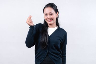 Siyah hırkalı gülümseyen bir Asyalı kadın, uzun siyah saçlı, tek eliyle bir parmak kalp hareketi yapıyor, tarafsız bir arkaplana karşı..