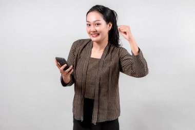 Geleneksel çizgili Lurik ya da Sorjan kıyafeti giyen, elinde akıllı telefon olan ve başarıyı kutlayan neşeli Endonezyalı bir kadın. Kültürel miras ve modern dijital yaşam tarzının karışımı..