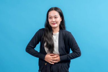 Genç Asyalı kadın iki eliyle karnını tutuyor, mavi arka planda dikiliyor, günlük kıyafetler giyiyor. Sağlık sorunları, karın ağrısı, sindirim veya rahatsızlık kavramı.