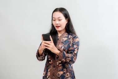 Çiçek desenli kıyafetli genç bir kadın, akıllı telefonunu kullanarak düz bir arkaplana karşı çekici bir ifade sergilerken şaşırmış görünüyor.