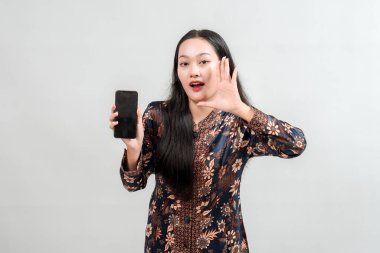 Geleneksel Batik gibi giyinmiş genç bir kadın elinde akıllı bir telefon ve jestle sanki heyecan verici haberleri paylaşıyormuş ya da cihazla ilgili bir duyuru yapıyormuş gibi hareket ediyor..