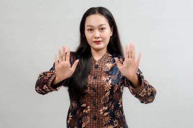 Geleneksel Batik gibi giyinmiş genç bir kadın, ellerini bir açıklık jesti ile kaldırdı. Tarafsız bir arkaplanda stüdyo resmi.