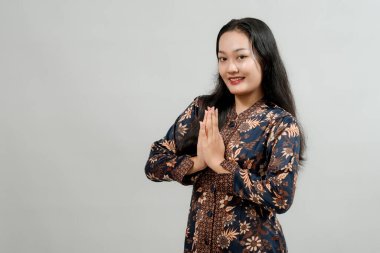 Batik gibi giyinmiş genç bir kadın, elleri göğsünde, şükranlarını sunuyor. Tarafsız bir arkaplanda stüdyo resmi.