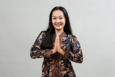 Batik gibi giyinmiş genç bir kadın, elleri göğsünde, şükranlarını sunuyor. Tarafsız bir arkaplanda stüdyo resmi.