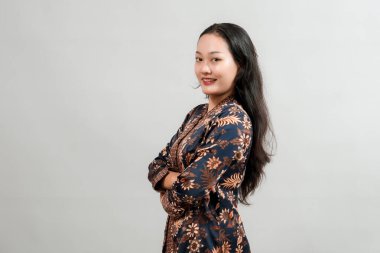 Geleneksel batik elbise giyen, kollarını kavuşturup gülümseyen, tarafsız bir arkaplanda poz veren kendinden emin genç Asyalı kadın..