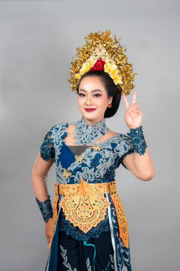 Modern Bali geleneksel kıyafeti giymiş Asyalı bir kadın yumuşak ve kendinden emin bir gülümsemeyle barış işareti gösteriyor. Çekici ifadesi ve şık görünüşü reklam amaçlı..