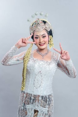 Geleneksel Sundan kıyafetleri ve süslenmiş taç giymiş güzel Endonezyalı gelin iki eliyle barış işareti yapıyor. Gülüşü, detaylı makyajı ve elbisesi zarafet ve neşe saçıyor..