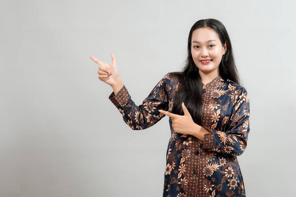 Молодая женщина в стильном индонезийском батике, уверенно указывая на копировальное пространство с дружелюбной улыбкой, идеально подходит для демонстрации продуктов или рекламных акций, связанных с индонезийской культурой
