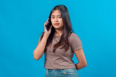 Kendine güvenen genç Asyalı bir kadın telefon görüşmesi yapıyor, parlak mavi bir arkaplan karşısında duruyor, ciddi görünüyor ve mobil iletişim sırasında dinlerken odaklanıyor..