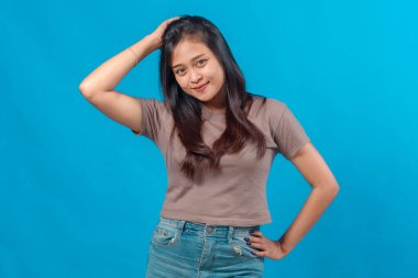 Uzun siyah saçlı gülümseyen genç Asyalı kadın, bir eliyle kafasına, diğer eliyle beline dokunarak, parlak mavi bir arkaplana karşı güven içinde poz veriyor..