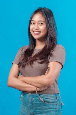 Kendine güvenen, uzun düz saçlı, kameraya gülümseyen, kollarını kavuşturmuş, günlük kıyafetler giyen, parlak mavi stüdyo arka planında soyutlanmış genç bir Asyalı kadının yakın plan portresi..