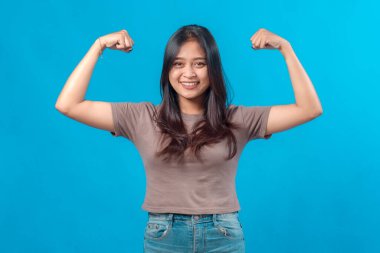 Kendine güvenen genç Asyalı kadın gülümserken iki kol kasını da güçlü bir şekilde gösteriyor. Mavi arkaplanda stüdyo portresi, güçlendirmeyi, sağlığı ve kararlılığı simgeliyor.