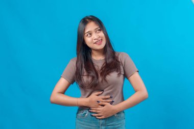Genç Asyalı bir kadın gülümseyerek karnına dokunuyor ve dilini şakayla uzatıyor. Aç bir ifadeyle yukarı bakıyor. Parlak mavi bir arka planda izole edilmiş..